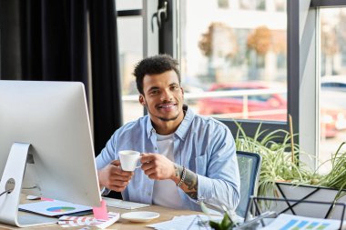 Smiling man sips coffee while working at a stylish desk in a bright workspace. clipart