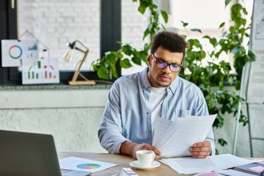 Concentrated man examining paperwork while sitting at a stylish desk with plants and charts. clipart