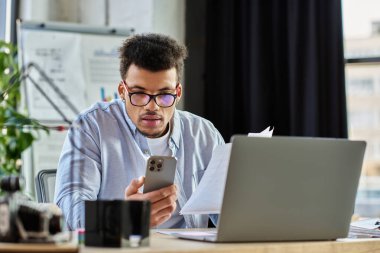 Genç adam şık ofis masasında doküman incelerken akıllı telefonunu kontrol ediyor..