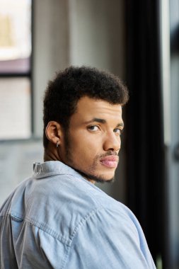 A stylish man with curly hair looks back with a contemplative expression in a bright room. clipart