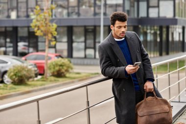 A fashionable man leans against a railing while using his smartphone in an urban setting. clipart