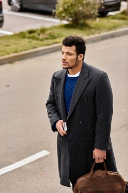 A handsome man with a confident expression strolls through a quiet parking lot holding a bag. clipart