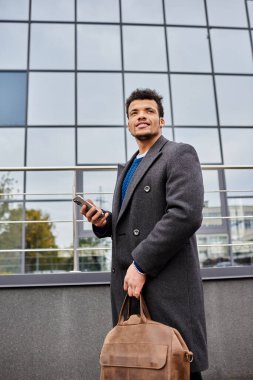 Yakışıklı bir adam şık bir binanın dışında duruyor, gülümsüyor, telefonunu ve çantasını tutuyor..