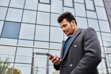 Modern bir ofis binasının önünde akıllı telefonunu kullanan şık bir adam gülümser..