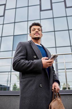 Şık ceketli neşeli bir adam telefonunu kontrol ederken çağdaş bir binanın önünde duruyor..