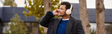 A charming man listens to music with headphones while strolling through a peaceful park setting. clipart