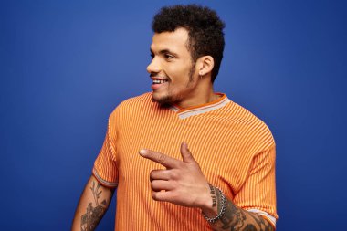 Dövmeleri olan şık bir adam turuncu çizgili bir gömlek giyerken gülümseyen neşeli hareketler yapar..