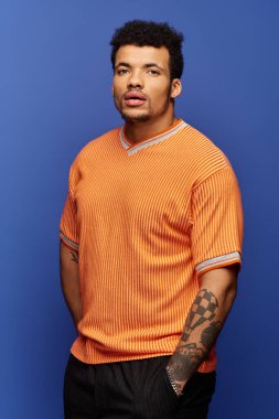 Bir adam göz alıcı turuncu gömleğiyle karizmasını sergiliyor, canlı mavi bir duvara karşı duruyor..