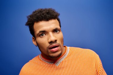 Büyüleyici bir adam mavi duvarın önünde turuncu çizgili süveter giyerken şaşırıyor..