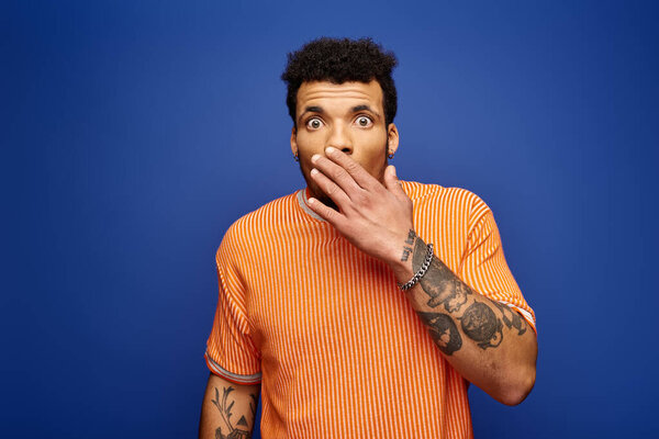 Красивый мужчина с татуировками выражает удивление широкими глазами и игривым жестом.