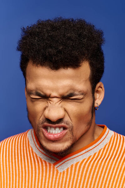 Стильный человек демонстрирует игривую гримасу с энтузиазмом, установленную на ярком синем фоне.