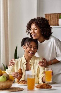 Two women enjoy breakfast together, smiling and sharing a warm moment in their apartment. clipart