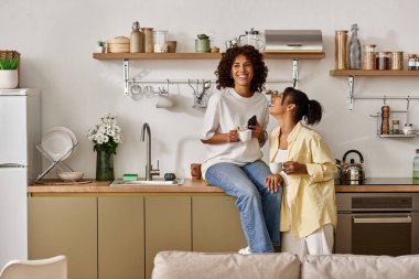 Two women share a joyful moment in their sleek apartment, sipping tea and laughing together. clipart