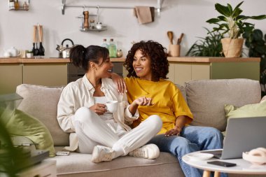 Two young women share laughter and coffee while chatting in their stylish apartment. clipart