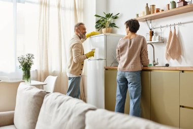 Olgun bir çift, güneşli bir öğleden sonra mutfağını temizler, gülümsemelerini ve kahkahalarını paylaşır..