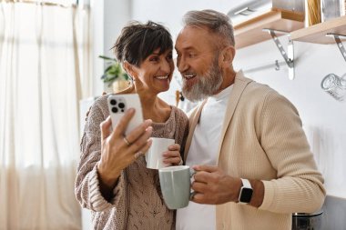 Yaşlı çift neşeli bir anı paylaşıyor, ellerinde fincanlarla güneşli bir mutfakta anılarını yakalıyorlar..
