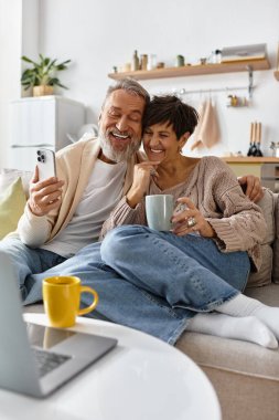 Senior man and woman enjoying a warm drink together while smiling at a smartphone. clipart