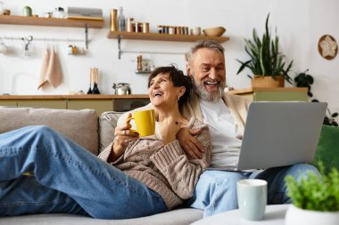 Neşeli, olgun bir çift kanepede oturur, dizüstü bilgisayar kullanırken kahkahalarını ve sıcak içkilerini paylaşırlar..