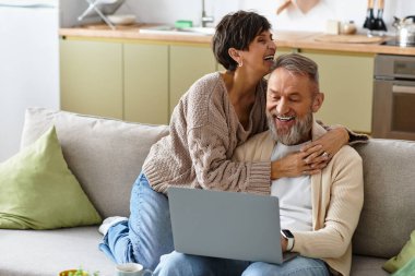 Couple laughs together on a cozy sofa while browsing online in their modern home. clipart