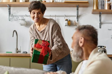 Mature woman presents a beautifully wrapped gift to her smiling partner in a cozy living space. clipart