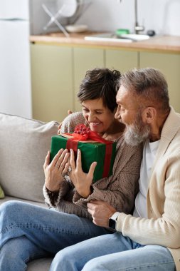 A mature couple smiles joyfully while unwrapping a thoughtful gift in their cozy living room. clipart