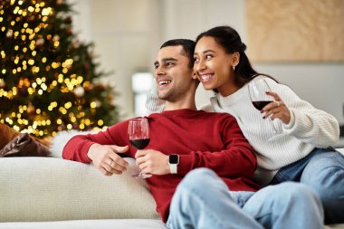 A couple sips wine together amid festive decor and a Christmas tree. clipart