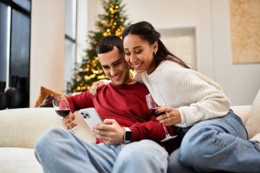 A beautiful couple shares laughter and wine while relaxing on a couch during the holidays. clipart