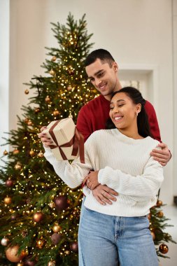 Couple joyfully sharing a moment together while unwrapping a gift under the Christmas tree. clipart