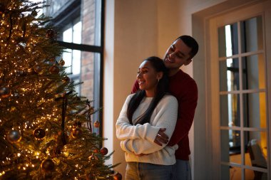 A beautiful couple shares a warm embrace by the shimmering Christmas tree, celebrating love. clipart