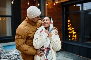 A couple enjoys each others warmth outdoors, surrounded by shimmering Christmas lights. clipart