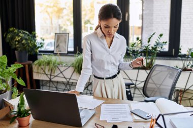 Focused young African American businesswoman reviews documents while standing at her desk. clipart