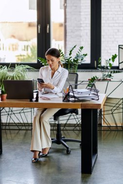 A young Black woman in a white shirt uses her smartphone at her desk in a bright office. clipart