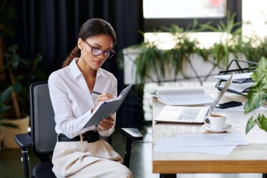 A young african american woman in a white shirt takes notes while seated at a modern office desk. clipart