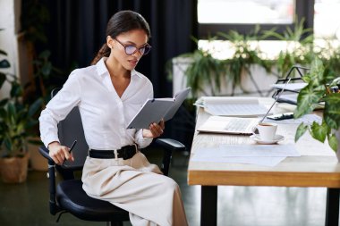 Young professional in a white shirt reviews documents while seated at her office desk. clipart