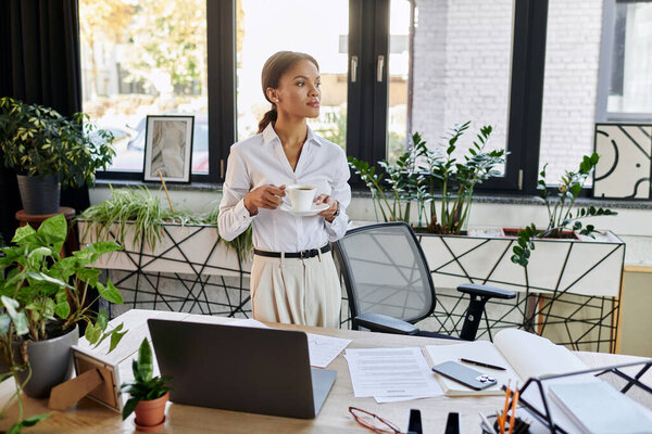 Профессиональная молодая женщина в белой рубашке стоит с чашкой кофе, отражая в своем рабочем пространстве.