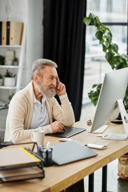 Mature man focused on computer at his workspace, surrounded by plants in a bright office. clipart