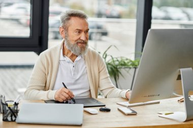 Sakallı kıdemli adam bitkilerle dolu geniş bir ofis masasında dijital işler yapıyor..