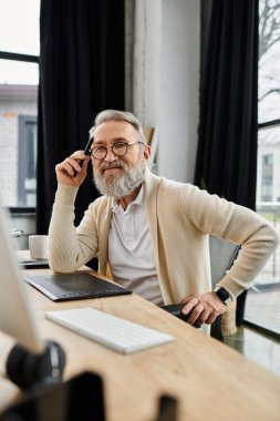 Mature man with glasses sits at a desk, reflecting while looking at a computer screen in an office. clipart