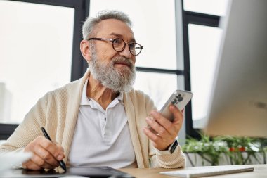 Yakışıklı son sınıf öğrencisi, parlak bir ofiste masa başında çalışırken akıllı telefonla iletişim kuruyor..