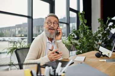 A mature handsome man engages in a phone conversation while sitting at a desk in a bright office. clipart