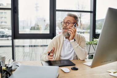 A mature and handsome man is engaged in a phone conversation while seated in a bright office. clipart