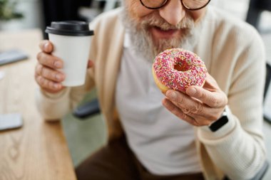 Sakallı, olgun bir adam elinde pembe bir çörek ve kahve tutarken gülümsüyor..