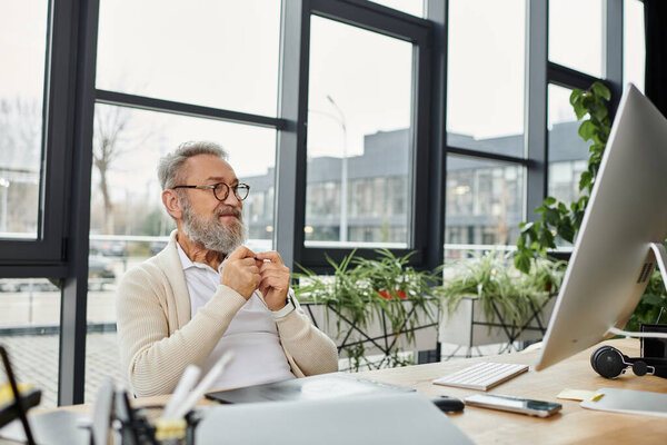 A handsome man works on a computer at a desk in a spacious, plant filled office.