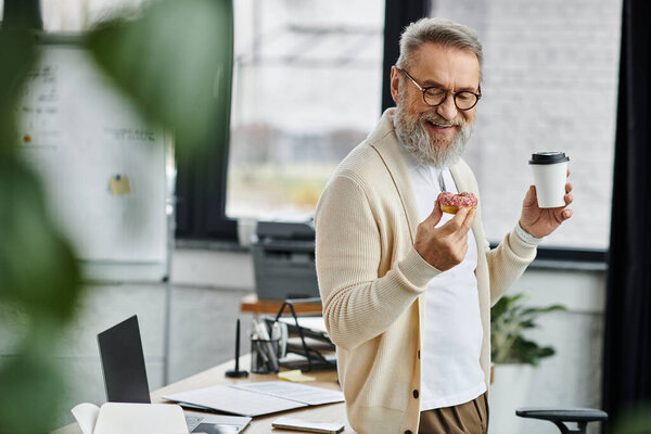 Handsome senior man shares a joyful moment with a pastry and coffee in a stylish workspace.