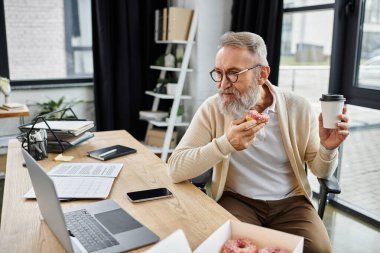 Handsome senior man relaxes with a donut and coffee while engaged with his laptop at work. clipart