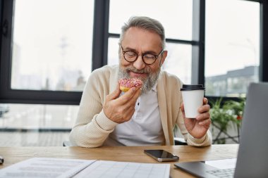 Elinde donut ve kahve tutarken gülümseyen gözlüklü olgun bir adam çalışma yerinde oturuyor..