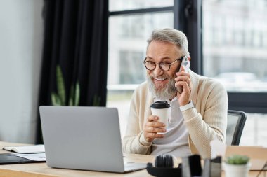 Olgun bir adam elinde kahve fincanı tutarken neşeli bir ifadeyle telefonda konuşur..
