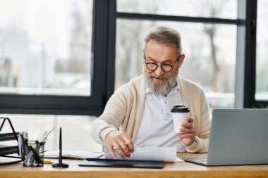 Olgun bir adam, elinde iyi aydınlatılmış bir ofis odasında kahve fincanı tutarken belgeleri gözden geçirir..