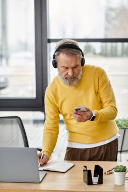 Handsome senior man wearing headphones engages with his smartphone at a desk. clipart