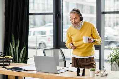 Sarı kazaklı adam çalışma odasında kahvesini yudumlarken bilgisayarla etkileşime geçiyor..